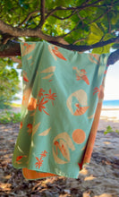 Light blue towel with beach-themed design hanging on a tree branch by a beach.